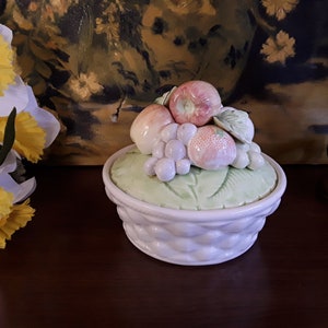 May include: A white ceramic fruit bowl with a lid. The lid is decorated with a 3D design of fruit, including apples, grapes, and a peach. The bowl has a woven design.