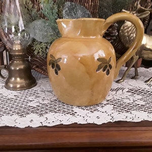 May include: A yellow ceramic pitcher with a brown glaze and olive branch designs. The pitcher has a wide mouth and a handle.