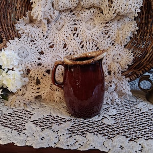 May include: A brown glazed ceramic pitcher with a handle. The pitcher is sitting on a white crocheted doily.