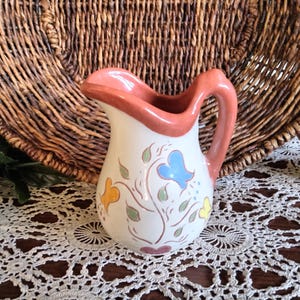 May include: A small, white ceramic pitcher with a peach-colored rim and handle. The pitcher is decorated with colorful floral designs in yellow, blue, and green. The pitcher is approximately 6 inches tall.