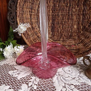 May include: A vintage pink glass basket with a clear, textured handle. The basket has a ruffled edge and sits on a white lace tablecloth. A woven basket and white flowers are in the background.
