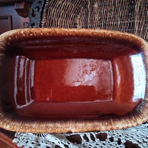 May include: A rectangular, ceramic serving dish with a glossy, reddish-brown interior and a textured, tan-colored rim. The dish is empty and sits on a white lace doily. The background includes a wooden surface and a woven basket.