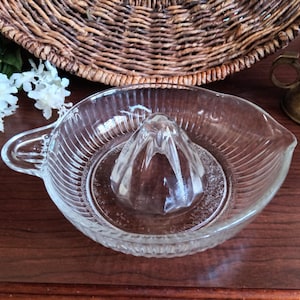 May include: A clear glass citrus juicer featuring a central reamer cone with faceted edges, a circular basin with vertical ribbed detailing along the outer walls, a small side handle, and a pouring spout on the opposite side of the handle.