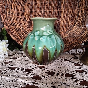 May include: A ceramic vase with a green and turquoise glaze, featuring a leaf pattern and brown vertical accents. The vase is displayed on a lace doily, with a wicker basket and a brass lamp in the background.