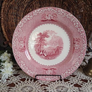 May include: A round ceramic plate featuring a pink and white transferware pattern. The center depicts a landscape with figures, while the rim is decorated with a continuous border of grapevines and small architectural vignettes in a matching pink tone.