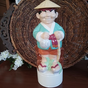 May include: A ceramic figurine of a smiling Asian man wearing a conical hat, teal jacket, and orange shorts. He holds a bag labeled "RICE" and a staff. The figurine stands on a white base, with red and white sandals. A decorative wicker basket is in the background.