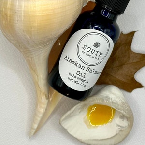 May include: A small glass bottle with a black dropper top and a white label. The label says "South of the Ocean" and "Alaskan Salmon Oil Wild caught. net wt. 1 oz". The bottle is sitting on a seashell.
