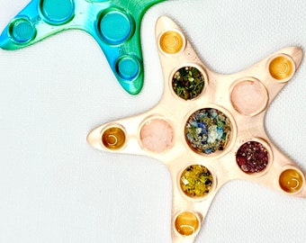 Könnte beinhalten: Zwei sternförmige Silikon-Fidget-Spielzeuge. Eines ist türkis und grün mit glänzender Oberfläche. Das andere ist hellbraun mit matter Oberfläche und hat in jedem der Kreise unterschiedlich farbige Glitzer eingebettet.