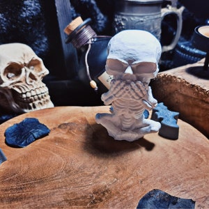 Może przedstawiać: Biała figurka szkieletu z rękami zakrywającymi usta, siedząca na drewnianej powierzchni z czaszką i innymi dekoracjami halloweenowymi.