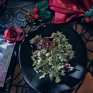 May include: A black plate containing a blend of dried herbs and flowers. The herbs are a mixture of green and brown, and the flowers are a deep red. The plate is on a lace tablecloth with a red satin fabric in the background.