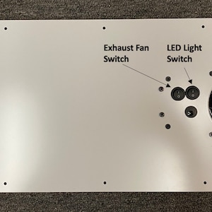 May include: White metal panel with two black toggle switches labeled "Exhaust Fan Switch" and "LED Light Switch". A black fan is mounted on the right side of the panel with the text "Exhaust Fan" below it.