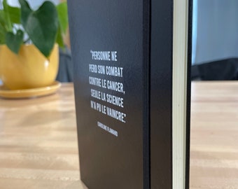 May include: A black notebook with a white quote on the cover that reads "PERSONNE NE PERD SON COMBAT CONTRE LE CANCER, SEULE LA SCIENCE N'A PU LE VAINCRE." The quote is attributed to CAROLINE FLAMAND.