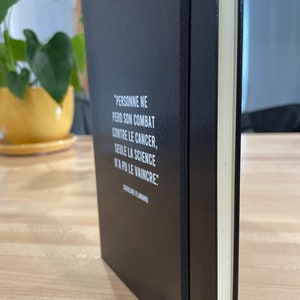 May include: A black notebook with a white quote on the cover that reads "PERSONNE NE PERD SON COMBAT CONTRE LE CANCER, SEULE LA SCIENCE N'A PU LE VAINCRE." The quote is attributed to CAROLINE FLAMAND.
