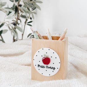 May include: A square wooden pencil holder featuring a circular white acrylic plaque on the front. The plaque has a black polka dot pattern, a red apple icon, and the text "Miss Honey" printed in black script. Several pens are inside the holder.