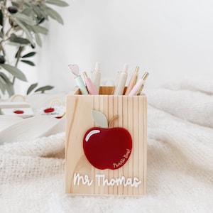 May include: A square wooden pencil holder featuring a red apple cutout with a light green leaf and the text Thank You. The name Mr Thomas is written in white script lettering below the apple. Several pens are stored inside the container.