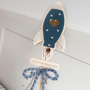 May include: Wooden rocket-shaped ornament with a blue body, white accents, and a gold heart. The ornament has a blue bow and the text "LOVED TO THE MOON & BACK". Decorative star and bead accents are attached.