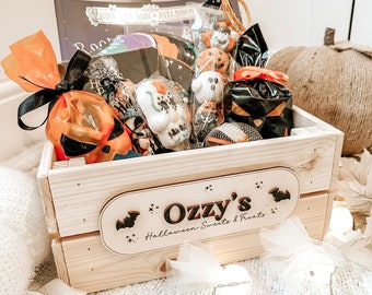 Puede incluir: Una caja de madera llena de golosinas de Halloween, incluyendo caramelos envueltos y un libro. La caja tiene un letrero de madera que dice "Ozzy's Halloween Sweets & Treats". La caja está decorada con un lazo negro y celofán naranja.
