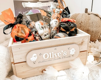 Puede incluir: Una caja de madera llena de golosinas y juguetes de Halloween. La caja está decorada con un letrero que dice "Olive's Halloween Sweets & Treats" y presenta una ilustración de un fantasma.