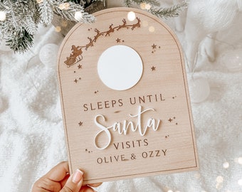 Puede incluir: Un letrero de madera con un círculo blanco en el centro, una silueta de Santa Claus en su trineo con renos, y el texto "Sleeps Until Santa Visits Olive & Ozzy".