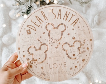 Könnte beinhalten: Ein rundes Holzschild mit Mickey Mouse-Motiv. Oben steht "Lieber Weihnachtsmann" und es enthält eine Liste von Weihnachtswünschen, darunter "Trink etwas, falls du Durst hast", "Ho Ho Ho", "Prost", "Probiere meinen Lieblingskeks", "Guten Appetit" und "Leckerlis für die Rentiere". Es ist signiert mit "Liebe, Olive & Ossy".