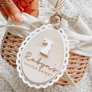 May include: A white wicker basket with a white fabric lining and a wooden tag that says "Robyn's Easter Basket" with a bunny silhouette. The basket has a brown ribbon tied around the handle.
