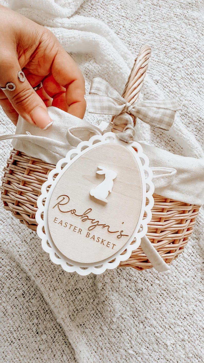 May include: A white wicker basket with a white fabric lining and a wooden tag that says "Robyn's Easter Basket" with a bunny silhouette. The basket has a brown ribbon tied around the handle.