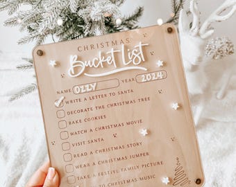 May include: A wooden Christmas bucket list sign with a white background and black text. The sign has a checklist of Christmas activities, including writing a letter to Santa, decorating the Christmas tree, baking cookies, watching a Christmas movie, visiting Santa, reading a Christmas story, wearing a Christmas jumper, taking a festive family picture, listening to Christmas music, and sending a Christmas card. The sign is personalized with the name "Ozzy" and the year "2024".
