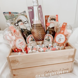 May include: A wooden crate filled with date night treats, including a bottle of sparkling wine, two bottles of beer, sweets, and marshmallows. The crate is labelled "Chelsea & Joe's date night treats."