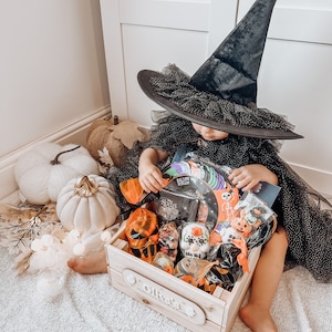 Puede incluir: Un ni&ntilde;o con un sombrero de bruja negro y un vestido est&aacute; sentado en una alfombra blanca y esponjosa. El ni&ntilde;o est&aacute; alcanzando una caja de madera llena de golosinas y juguetes con tem&aacute;tica de Halloween. La caja tiene un letrero personalizado que dice "Oliver's Halloween Treats & Fun".