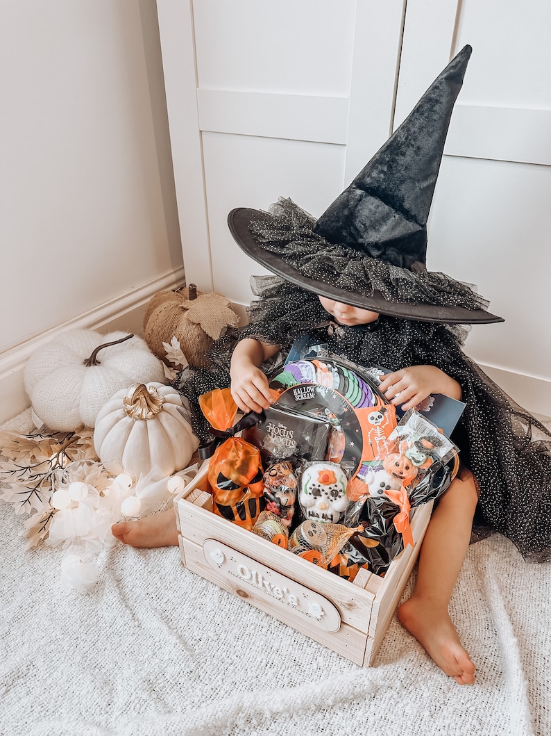 Puede incluir: Un ni&ntilde;o con un sombrero de bruja negro y un vestido est&aacute; sentado en una alfombra blanca y esponjosa. El ni&ntilde;o est&aacute; alcanzando una caja de madera llena de golosinas y juguetes con tem&aacute;tica de Halloween. La caja tiene un letrero personalizado que dice "Oliver's Halloween Treats & Fun".