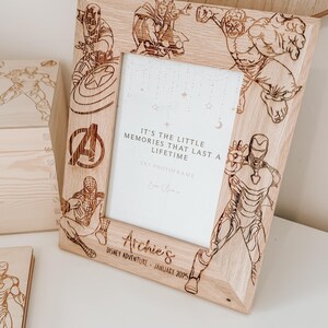Op de afbeelding: Een houten fotolijst met een lichte houtkleur. De lijst is versierd met afbeeldingen van Marvel-superhelden, waaronder Iron Man, Captain America, Thor, Hulk en Spiderman. De lijst heeft een witte passe-partout en een heldere glazen voorkant. De tekst "It's the little memories that last a lifetime" is gegraveerd op de lijst, samen met de tekst "5x7 Photoframe" en "Love Always". De lijst heeft ook de tekst "Archie's Disney Adventure - January 2025" gegraveerd op de onderkant.