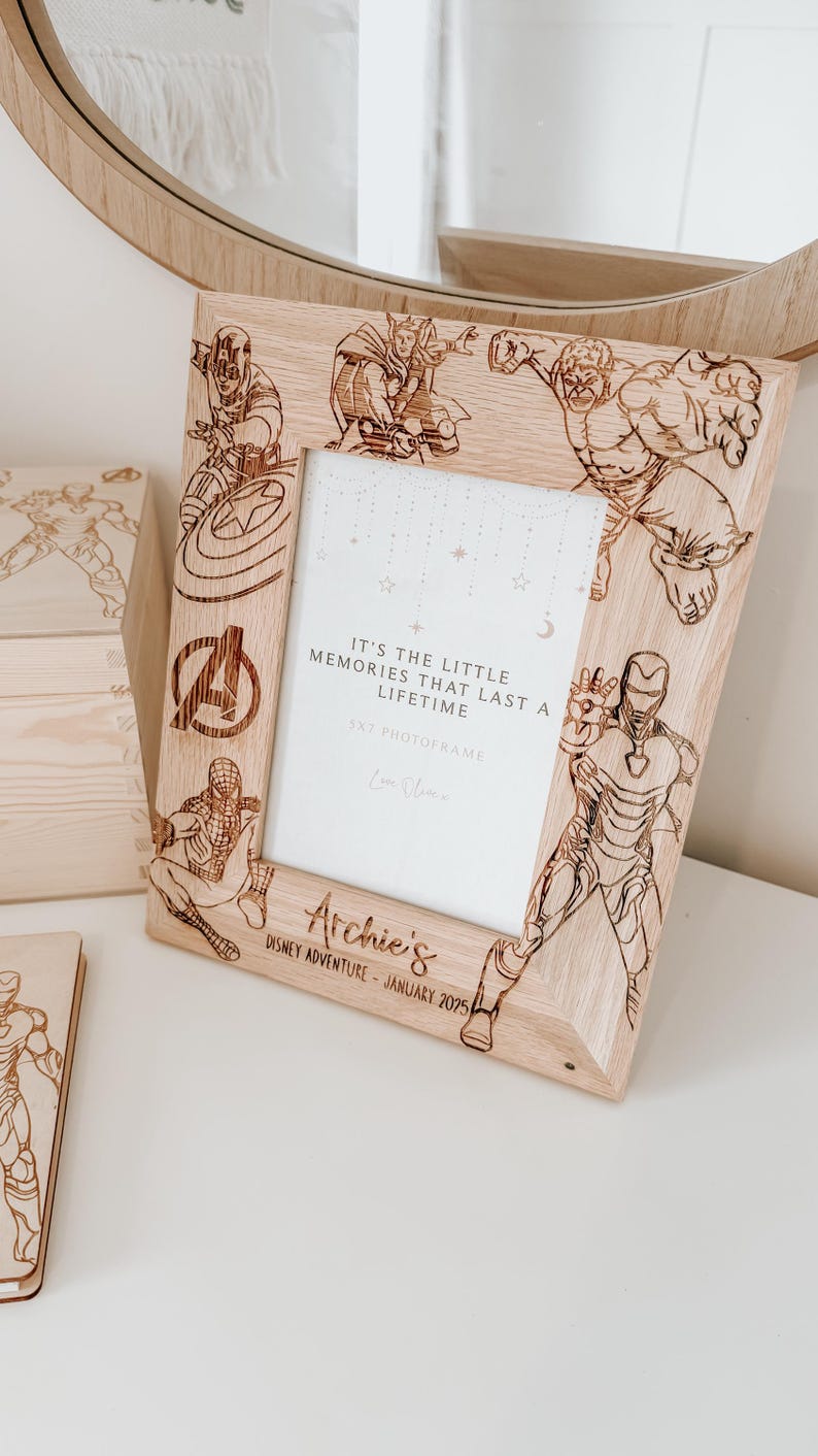 Op de afbeelding: Een houten fotolijst met een lichte houtkleur. De lijst is versierd met afbeeldingen van Marvel-superhelden, waaronder Iron Man, Captain America, Thor, Hulk en Spiderman. De lijst heeft een witte passe-partout en een heldere glazen voorkant. De tekst "It's the little memories that last a lifetime" is gegraveerd op de lijst, samen met de tekst "5x7 Photoframe" en "Love Always". De lijst heeft ook de tekst "Archie's Disney Adventure - January 2025" gegraveerd op de onderkant.