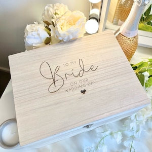 May include: A wooden box with a hinged lid, engraved with the words "To My Bride On Our Wedding Day" and a small heart. The box is light brown and has a natural wood grain.