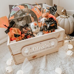 Puede incluir: Una caja de madera llena de golosinas de Halloween, incluyendo dulces, juguetes y decoraciones. La caja est&aacute; decorada con un fantasma y las palabras "Olive's Halloween Sweets & Treats".