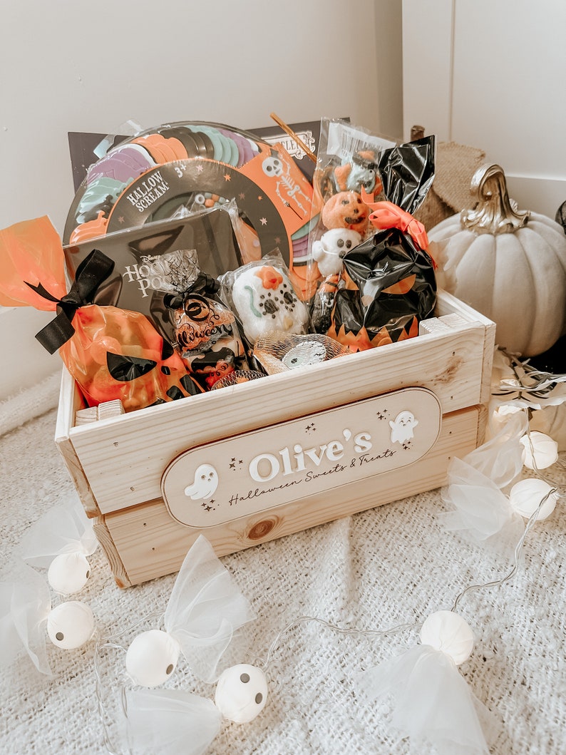 Puede incluir: Una caja de madera llena de golosinas de Halloween, incluyendo dulces, juguetes y decoraciones. La caja est&aacute; decorada con un fantasma y las palabras "Olive's Halloween Sweets & Treats".