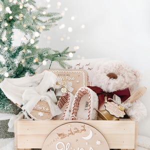 May include: A wooden crate labeled Olive's Christmas Eve Box with a festive scene, containing a plush teddy bear with a maroon bow, white slippers, a white garment, and a small wooden sign reading Official North Pole.