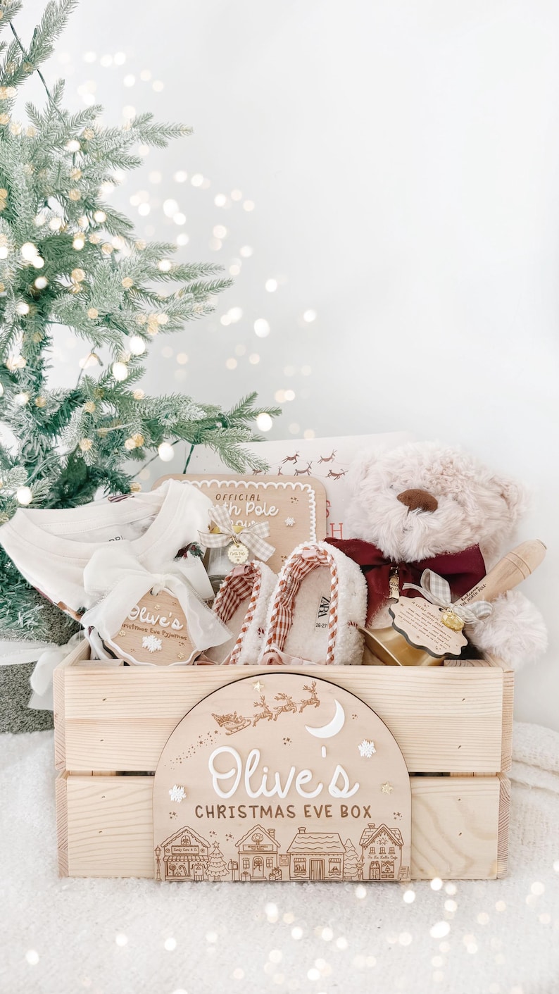 May include: A wooden crate labeled Olive's Christmas Eve Box with a festive scene, containing a plush teddy bear with a maroon bow, white slippers, a white garment, and a small wooden sign reading Official North Pole.