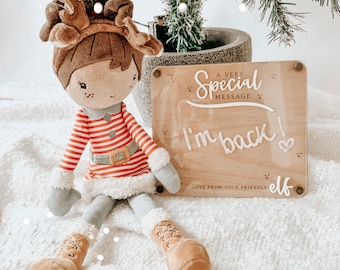 May include: A plush elf doll wearing a red and white striped dress with a brown headband and antlers. The doll is holding a wooden sign that says "A Very Special Message I'm Back! Love From Your Friendly Elf".