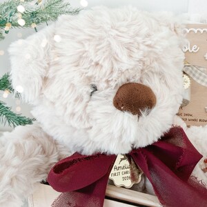 May include: A cream-colored plush teddy bear with a dark brown nose, wearing a deep burgundy sheer ribbon tied in a bow around its neck. A gold-toned metal tag hangs from the ribbon, engraved with the text Amelia FIRST CHRISTMAS 2026.