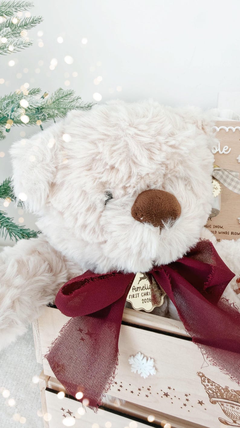 May include: A cream-colored plush teddy bear with a dark brown nose, wearing a deep burgundy sheer ribbon tied in a bow around its neck. A gold-toned metal tag hangs from the ribbon, engraved with the text Amelia FIRST CHRISTMAS 2026.