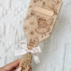 May include: Wooden hairbrush with engraved floral and fairy design. Features a light pink ribbon and the name "Florence" engraved on a small plaque. The brush has a natural wood finish and is decorated with flowers, butterflies, and a bird.