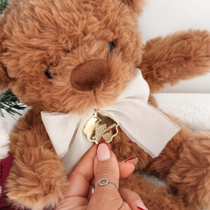 May include: A brown plush teddy bear with a cream-colored ribbon tied around its neck. Attached to the ribbon is a small, gold-colored, scalloped-edge metal tag engraved with the text Merry Christmas 2016 and a reindeer silhouette.