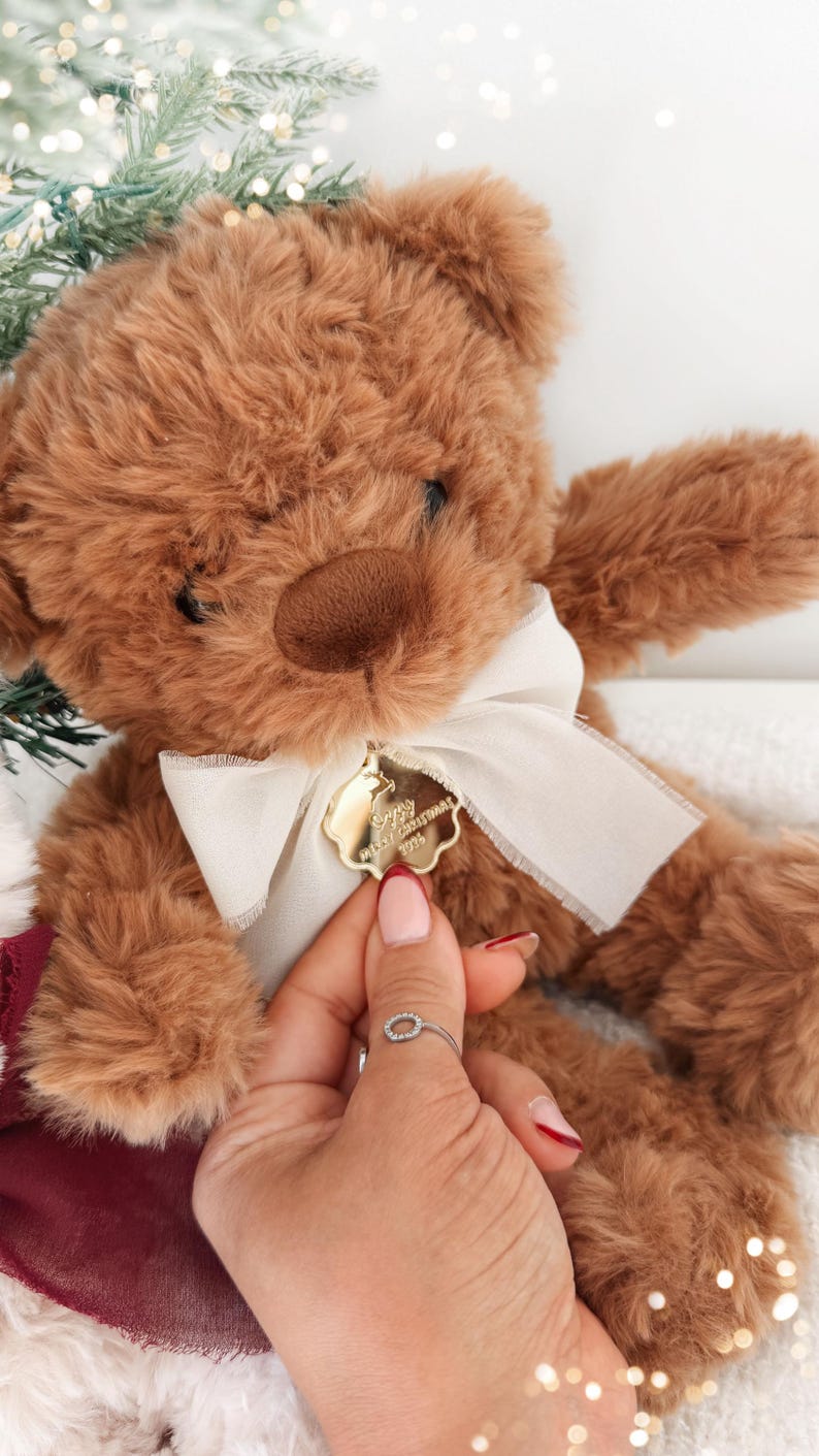 May include: A brown plush teddy bear with a cream-colored ribbon tied around its neck. Attached to the ribbon is a small, gold-colored, scalloped-edge metal tag engraved with the text Merry Christmas 2016 and a reindeer silhouette.