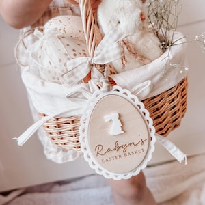 May include: A white wicker basket with a white fabric liner and a wooden tag that says "Robyn's Easter Basket" with a bunny silhouette. The basket is filled with a white stuffed bunny, white tissue paper, and white flowers.