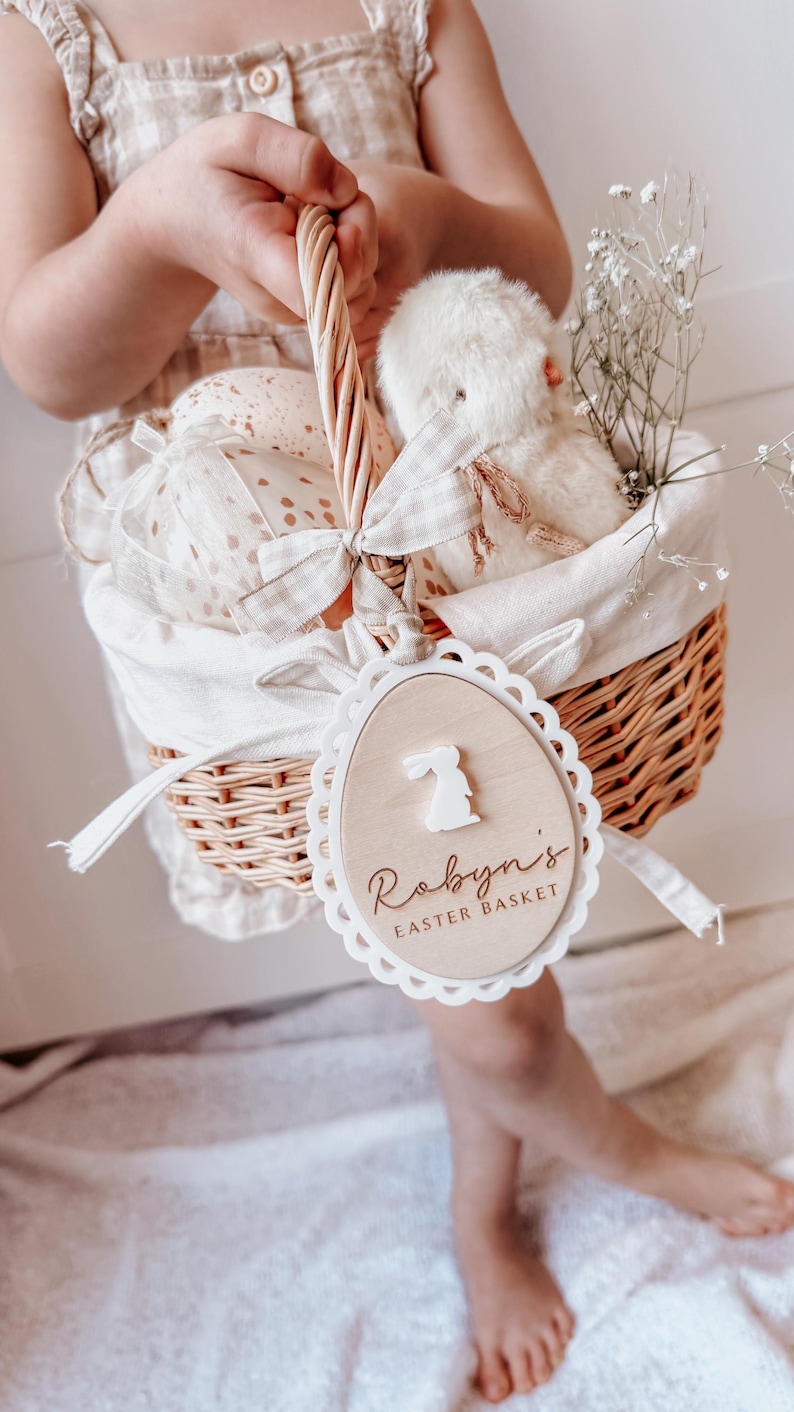 May include: A white wicker basket with a white fabric liner and a wooden tag that says "Robyn's Easter Basket" with a bunny silhouette. The basket is filled with a white stuffed bunny, white tissue paper, and white flowers.