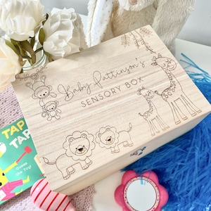 May include: A wooden sensory box with a light wood finish. The box has a lid and is decorated with a giraffe, lion, and monkey illustrations. The text "Baby Pattison's Sensory Box" is engraved on the lid.