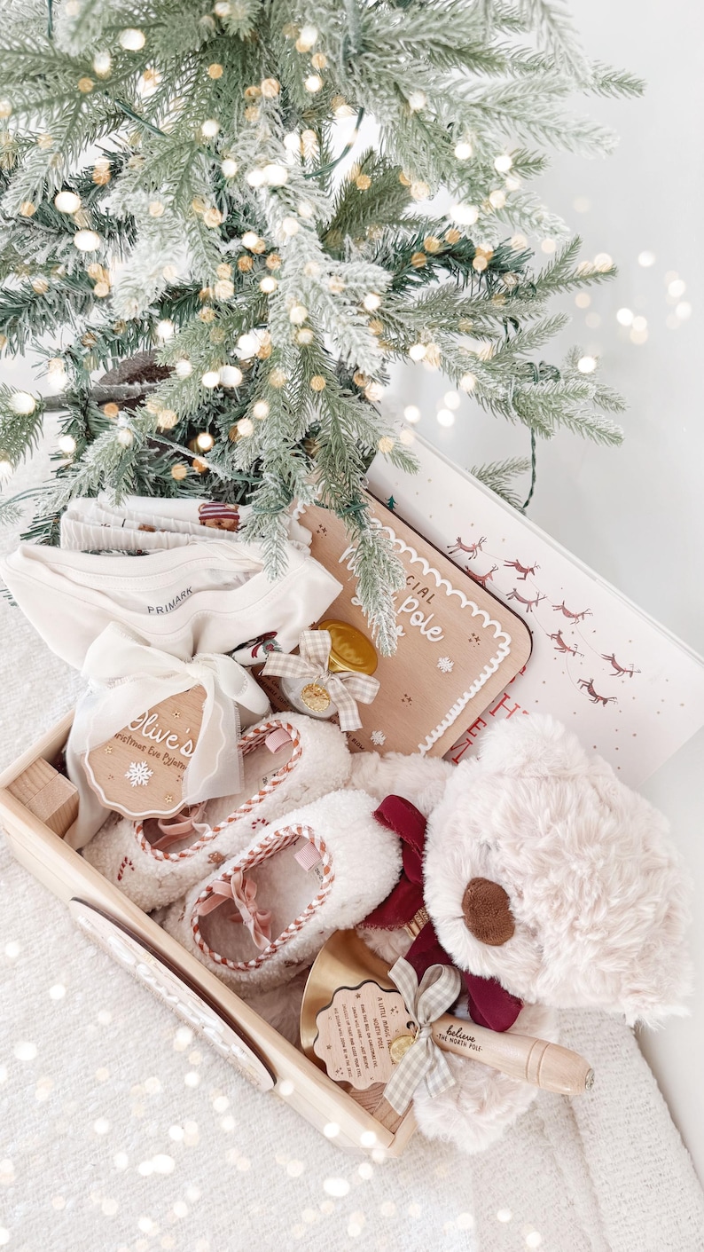 May include: A wooden gift crate containing a cream-colored teddy bear with a burgundy bow, fuzzy slippers with a candy cane pattern, a white long-sleeved shirt, a wooden sign reading Special North Pole Delivery, and a bell with a tag reading Believe.