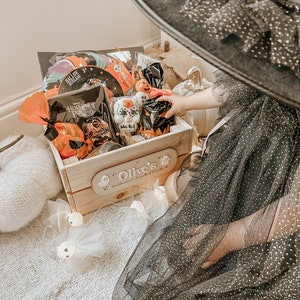 Puede incluir: Una caja de madera llena de golosinas y juguetes de Halloween. La caja est&aacute; decorada con un letrero personalizado que dice "Olive's Halloween Sweets & Treats". Un ni&ntilde;o con un vestido de tul negro y un sombrero de bruja est&aacute; alcanzando dentro de la caja.