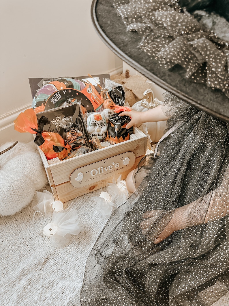 Puede incluir: Una caja de madera llena de golosinas y juguetes de Halloween. La caja est&aacute; decorada con un letrero personalizado que dice "Olive's Halloween Sweets & Treats". Un ni&ntilde;o con un vestido de tul negro y un sombrero de bruja est&aacute; alcanzando dentro de la caja.
