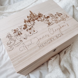 May include: A light-colored wooden memory box with a hinged lid. The box features an engraved design of a castle and cartoon characters, including Mickey Mouse, Minnie Mouse, and Donald Duck. The text "Axel James Claydon's MEMORY BOX" is also engraved on the lid.